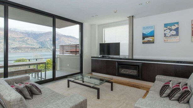 A modern living room with a glass coffee table, grey sofas, a flat-screen TV, and a fireplace. - Lakeshore Springs Apartments (Wanaka)