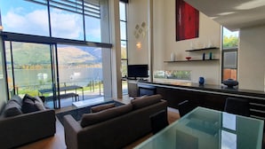A modern living room with a glass table and sofas, a kitchen with a large window, and a view of a lake and mountains. - Lakeshore Springs Apartments (Wanaka)