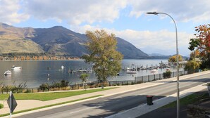 A lakeside road with a street lamp, a clear view of boats on the water, and mountains in the background. - Lakeshore Springs Apartments (Wanaka)