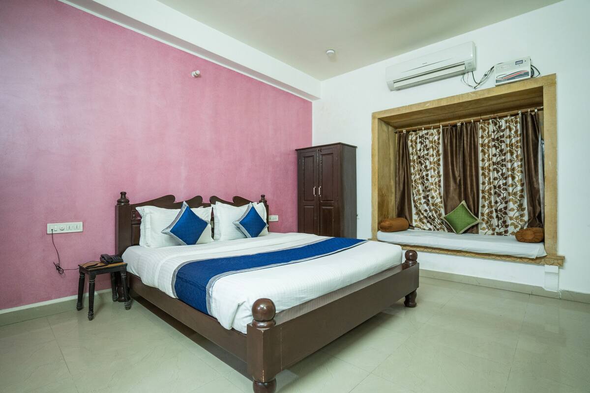 A bedroom with a wooden bed, pink walls, a window with patterned curtains, and an air conditioning unit.
