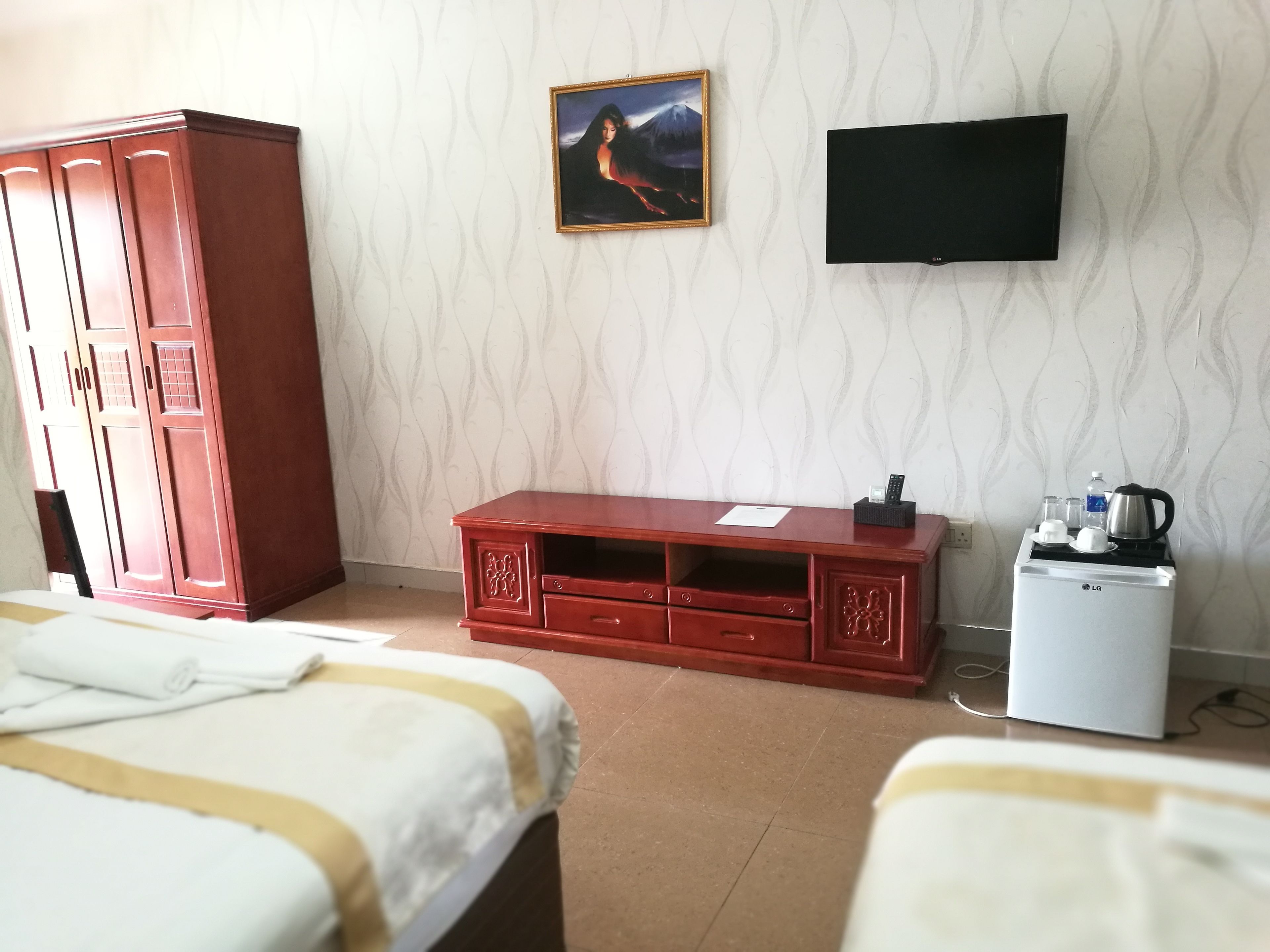 A hotel room with a wooden cabinet, a TV mounted on the wall, a small kitchenette with an LG fridge, and two beds with white and yellow bedding.