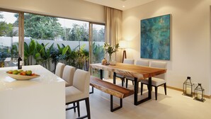 A modern dining area with a wooden table, white chairs, and a bench. There is a bowl of fruit on the table, a vase with flowers, and a painting on the wall. - Trichada Villas by RESAVA (Choeng Thale)
