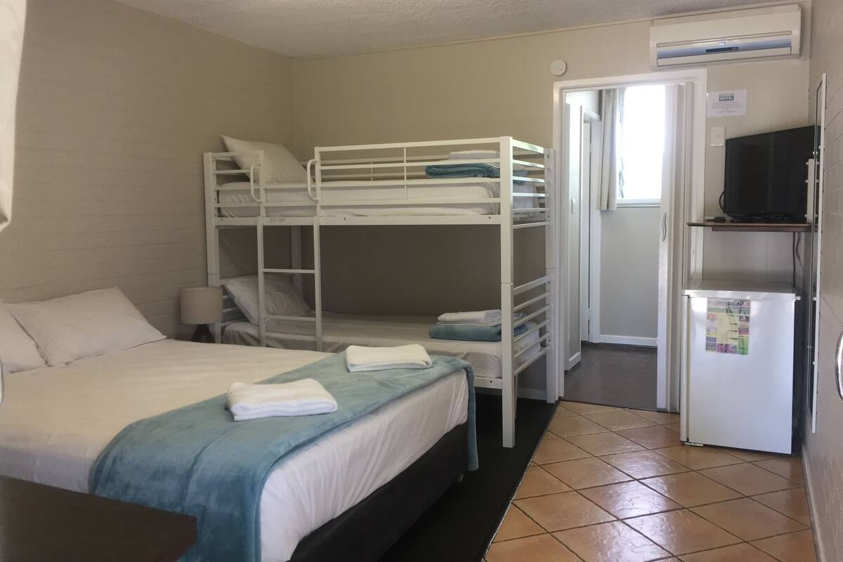 Quarto com beliche, escrivaninha, geladeira e porta para o banheiro.