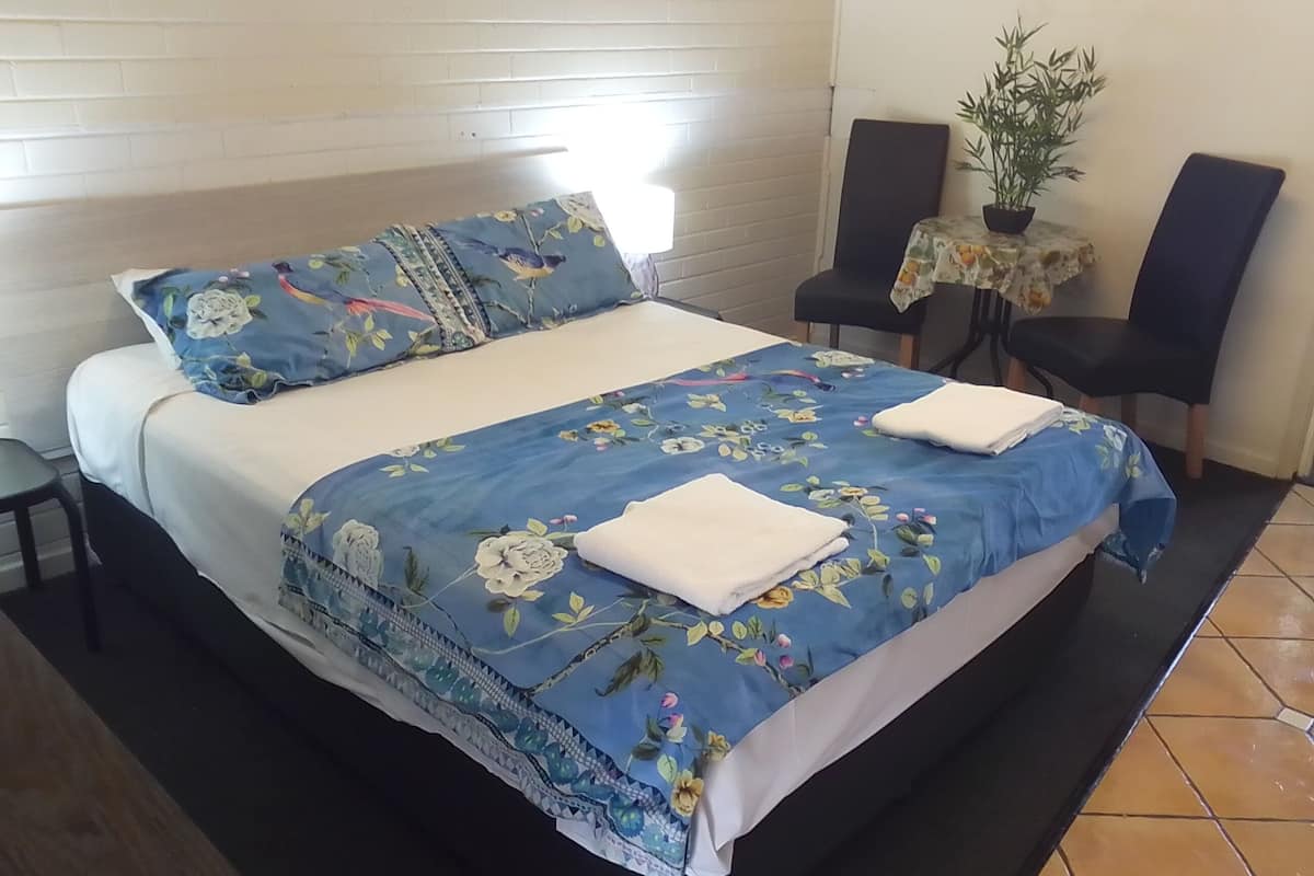 Cama bem arrumada com roupa de cama e almofadas florais, uma pequena mesa com uma planta e duas cadeiras num quarto com paredes de tijolo branco.