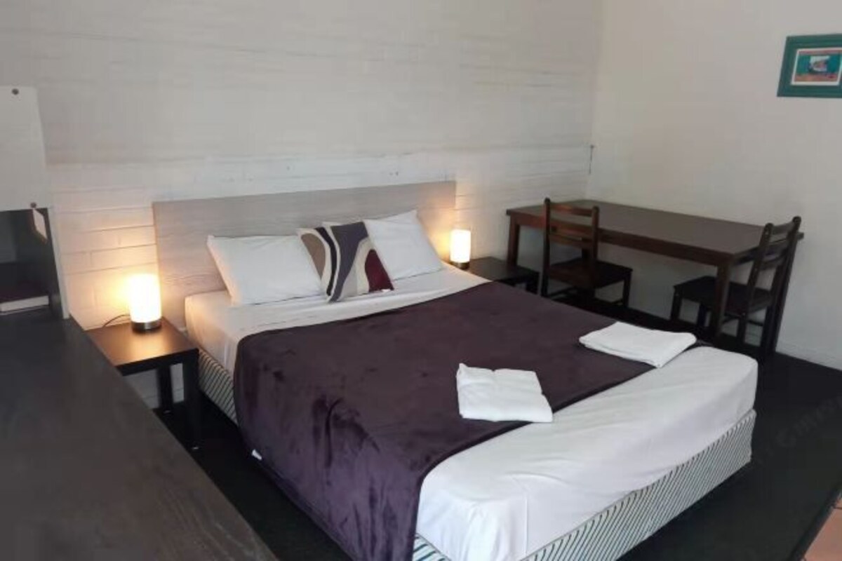 A neatly made bed with a purple blanket, white pillows, and a white duvet. There is a wooden nightstand with a lamp on it to the left of the bed. A wooden table with chairs is positioned to the right of the bed.