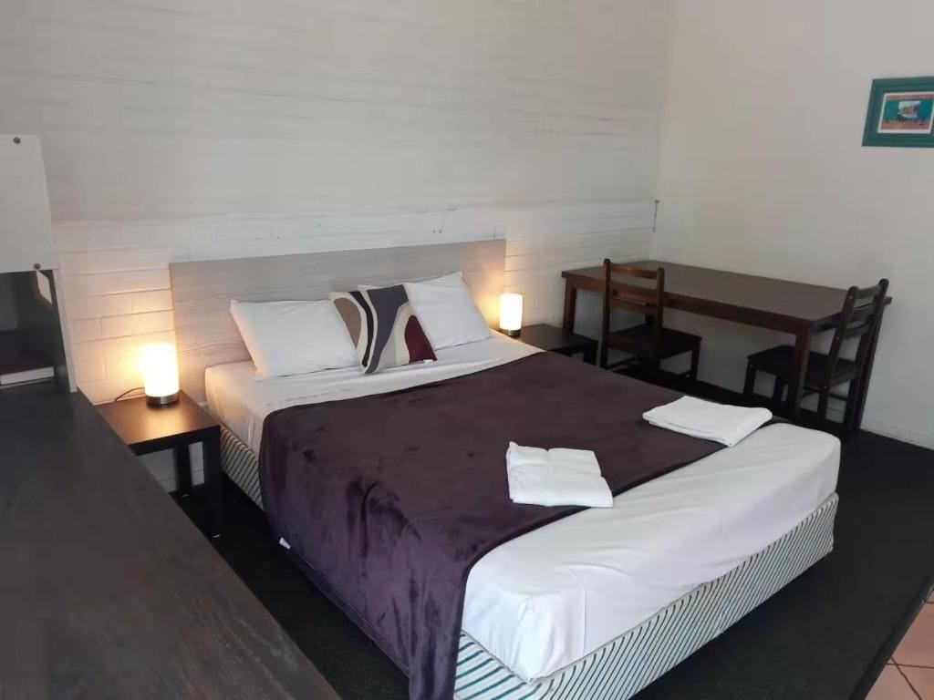 Una cama bien hecha con una manta morada y ropa de cama blanca, una mesita de noche con una lámpara, una silla de madera y una pequeña mesa con una silla.