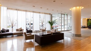 Un vestíbulo amplio con diseño moderno, que cuenta con una zona de estar grande con sofás y sillones, una mesa de centro y ventanas amplias que ofrecen vistas a la calle.