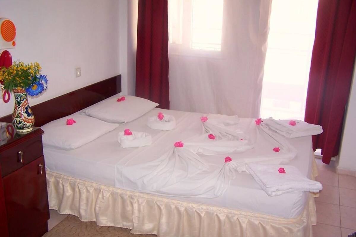 A neatly made bed with white linens and pink flower petals, a vase with flowers, and a red cabinet.