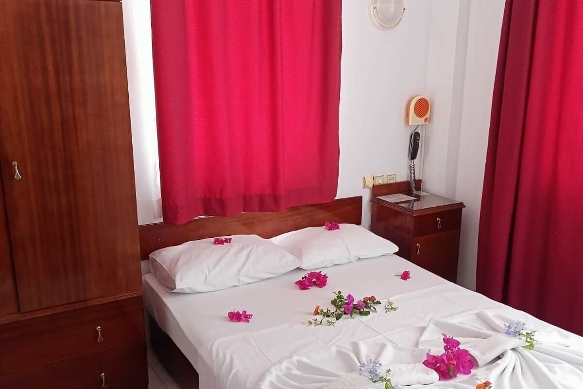 A hotel room with a bed, red curtains, a wooden wardrobe, a bedside table with a lamp, and a small round table with a map.