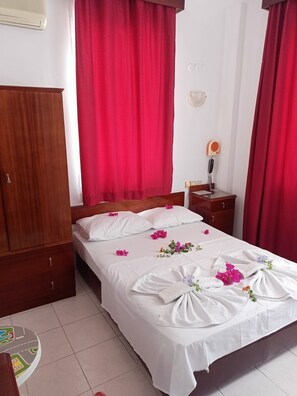 Quarto de hotel com cama, cortinas vermelhas, guarda-roupa de madeira, mesa de cabeceira com abajur e uma pequena mesa redonda com um mapa.