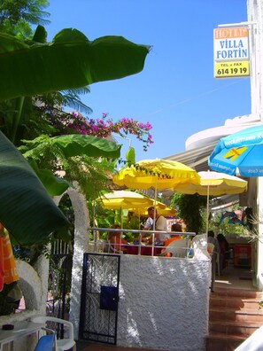 Um hotel chamado Villa Fortín com área externa e mesas sob guarda-sóis amarelos, cercado por vegetação exuberante e um céu azul límpido.