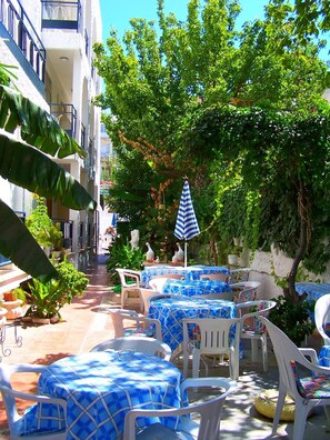 An outdoor seating area with tables and chairs, surrounded by greenery and a building with balconies. - Villa Fortin Pansiyon (Kusadasi)