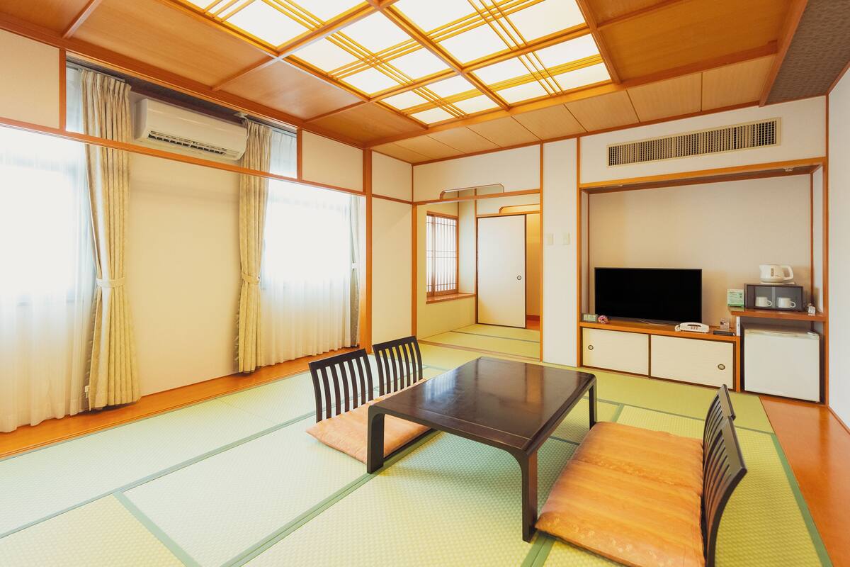 A traditional Japanese room with tatami flooring, a low table, chairs, a television, and a small kitchenette.