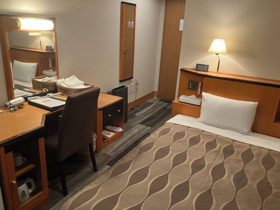 A hotel room with a bed, a desk, a chair, a lamp, a nightstand, a mirror, and a small clock.