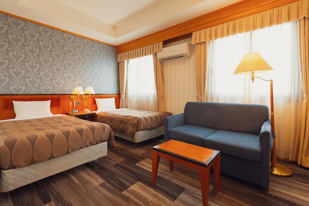 A hotel room with two beds, a sofa, a small table, an air conditioning unit, and a lamp.