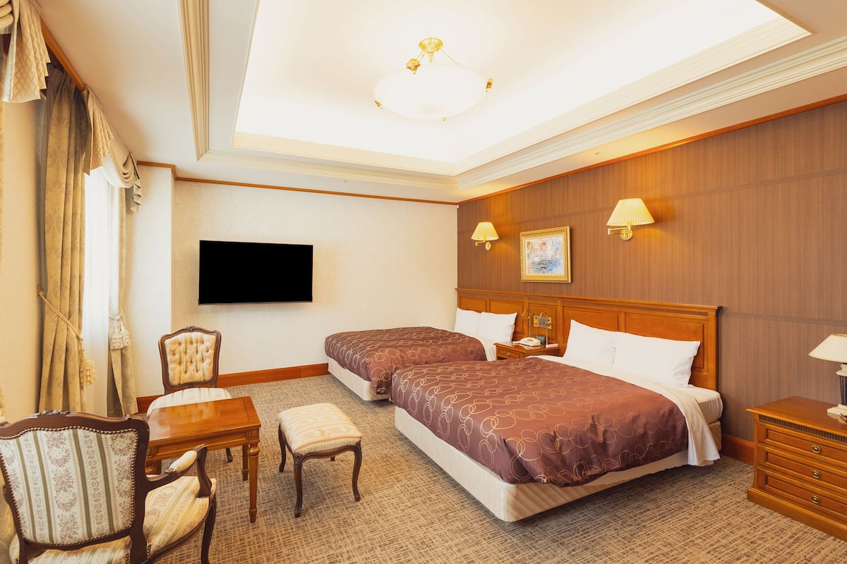 A hotel room with two beds, a desk, a chair, a television, and a ceiling light.