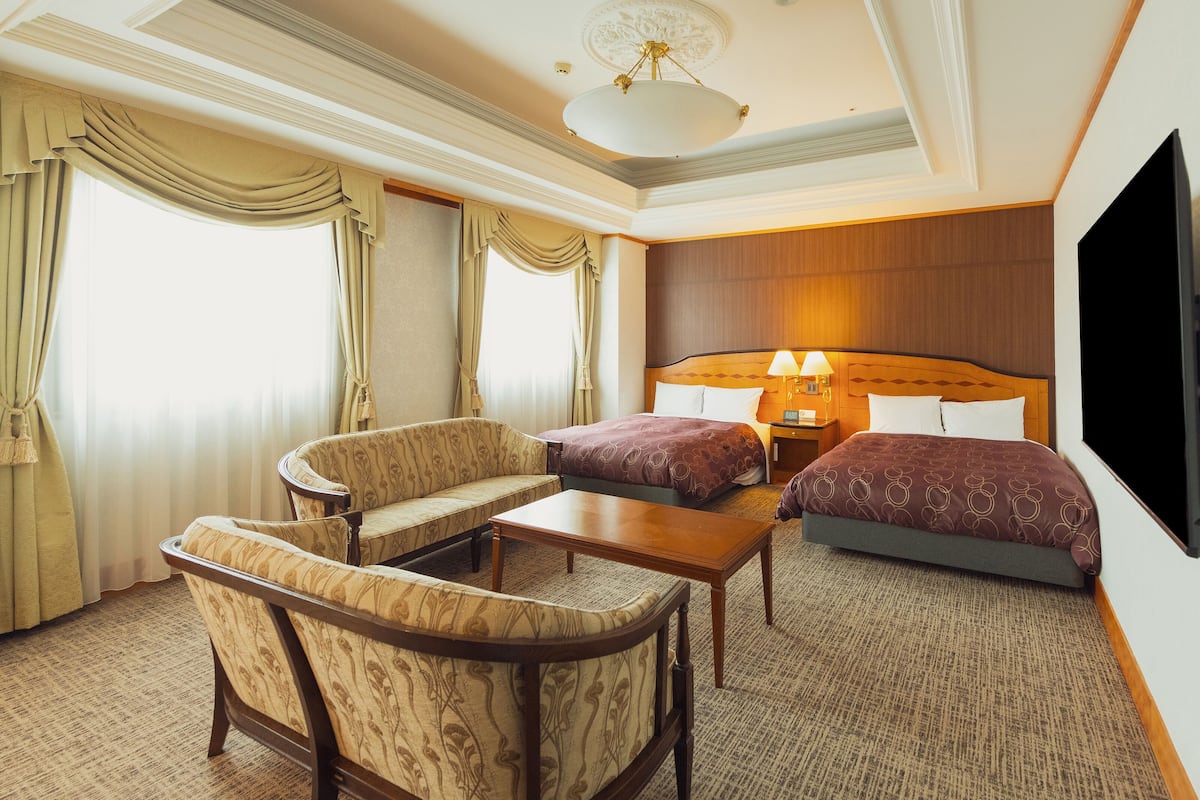 A hotel room with a sofa, two beds, a TV, a desk, and a chandelier.