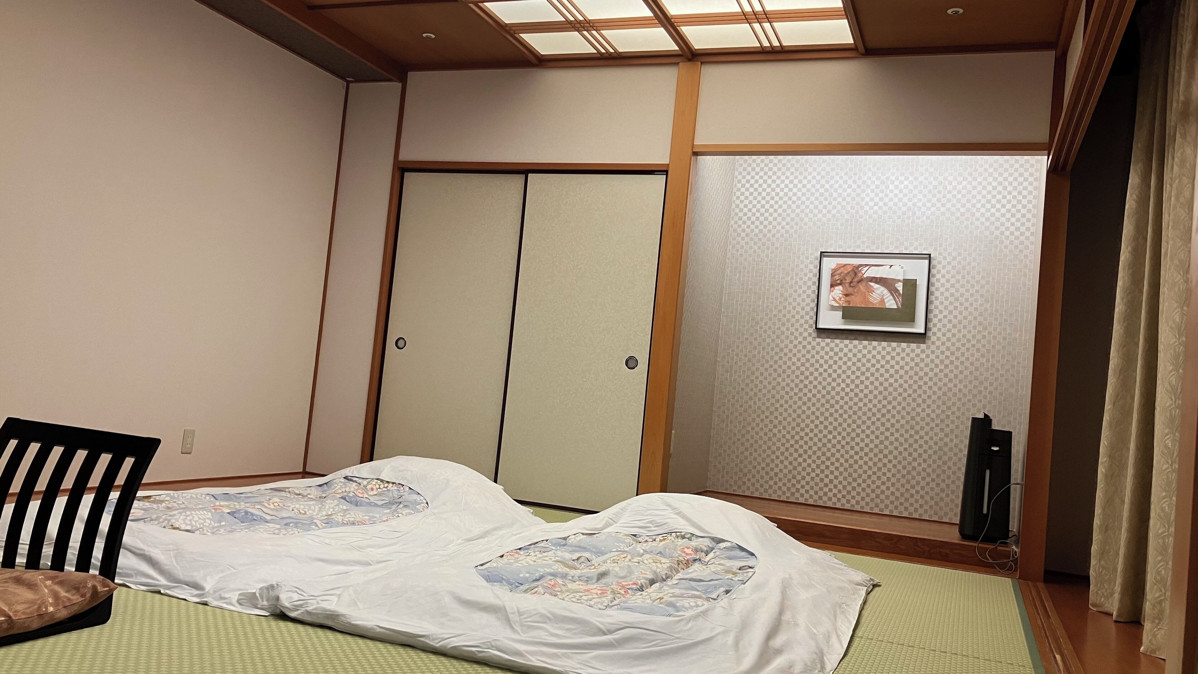 A room with tatami mats, a bed with a floral bedspread, a black chair, a sliding door, and a framed picture on the wall.
