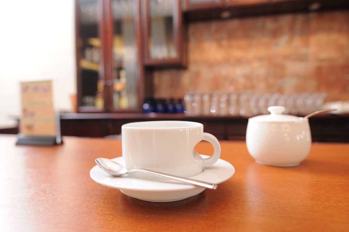A white cup and saucer with a spoon on a wooden table, a white teapot, and a blurred background with a menu stand.