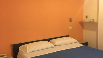 Hotelkamer met een oranje muur, een bed met witte kussens en een blauwe spreidek, een nachtkastje met een lamp en een klein plankje met een bordje.
