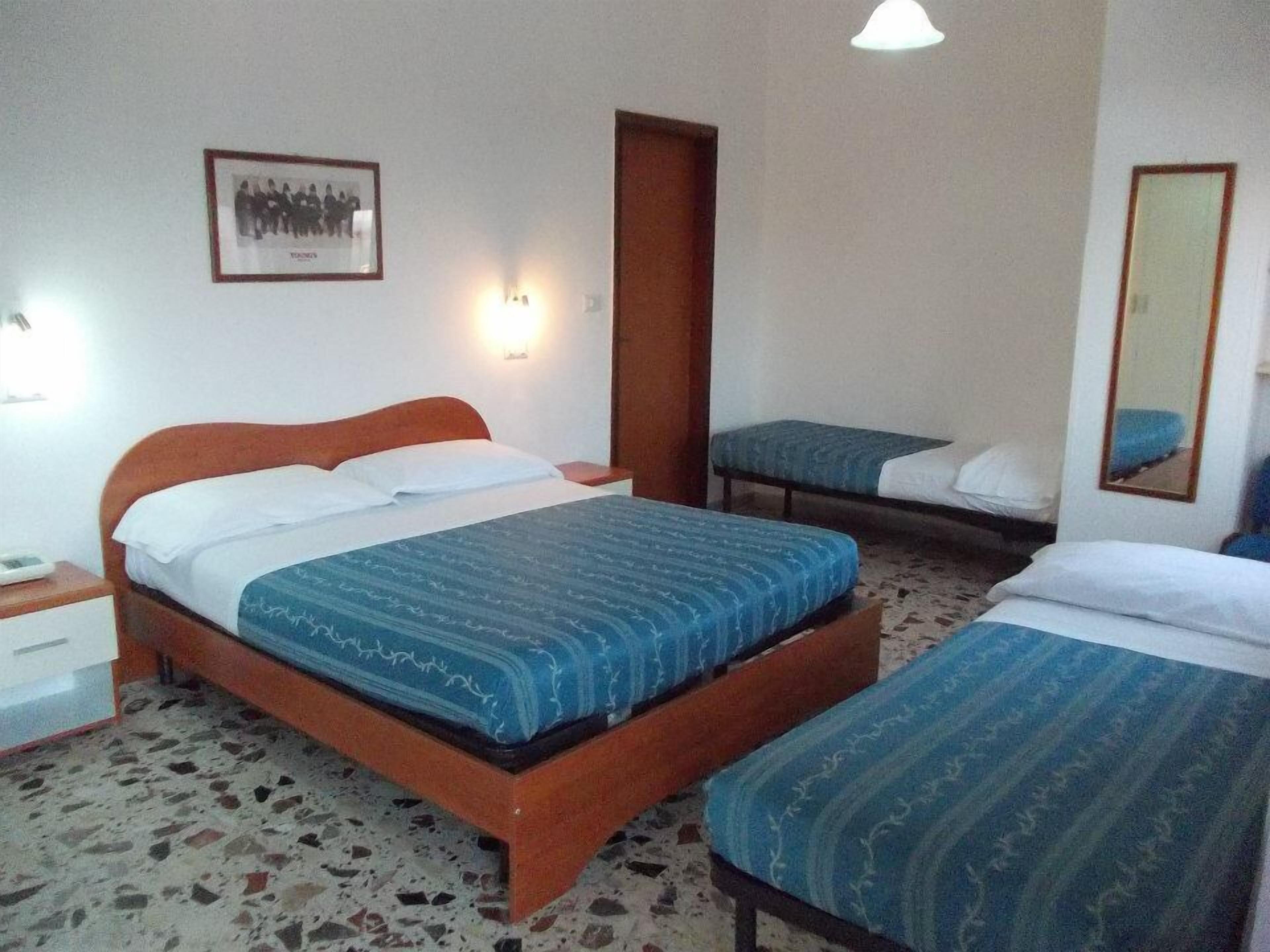 Ein Hotelzimmer mit einem großen Bett, einem kleineren Bett, einem Nachttisch, einer Wandleuchte, einem gerahmten Bild und einem Spiegel.