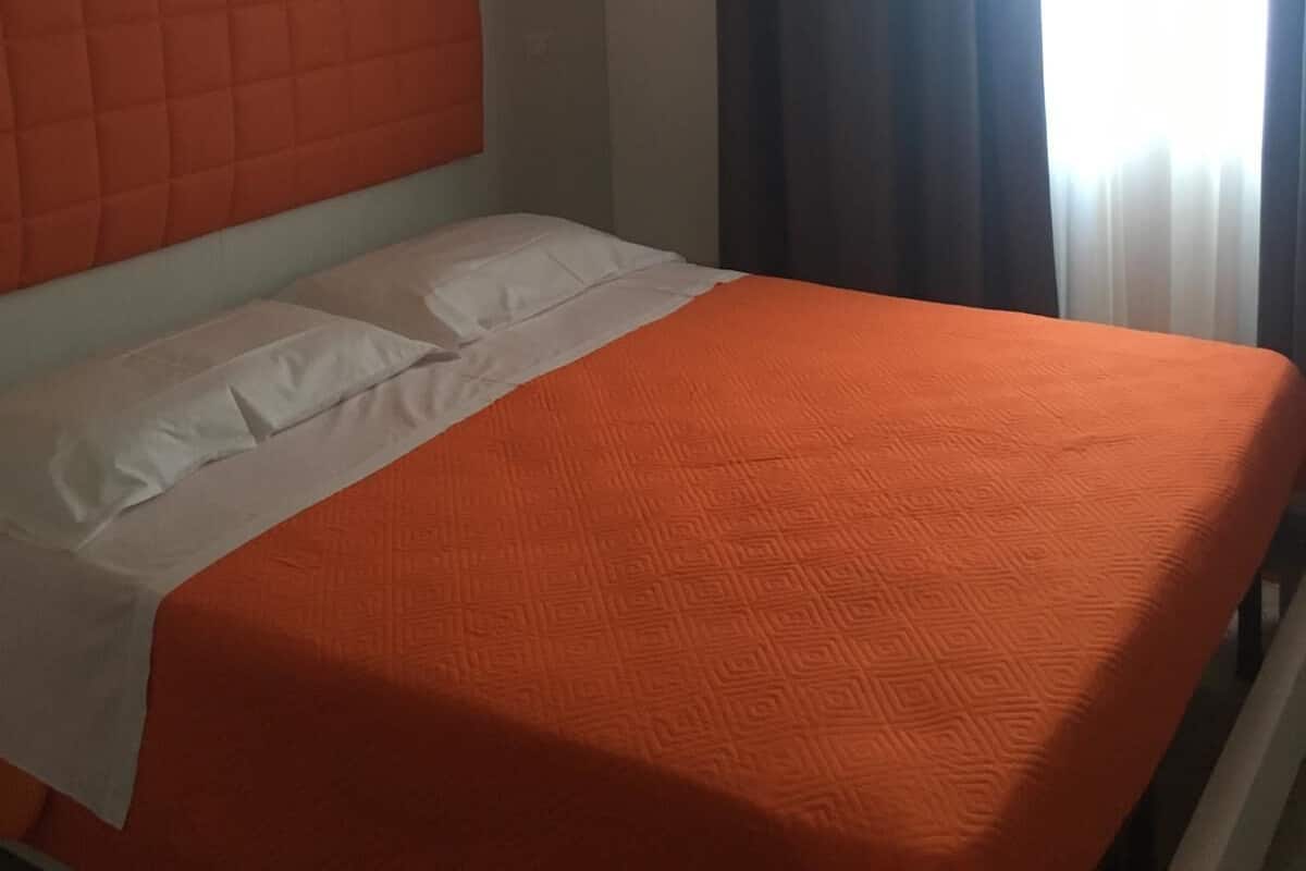 A hotel room with a bed, white headboard, orange bedspread, and a window with curtains.