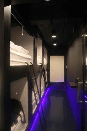 A narrow corridor with bunk beds on one side, each with a headboard and a hanging light.