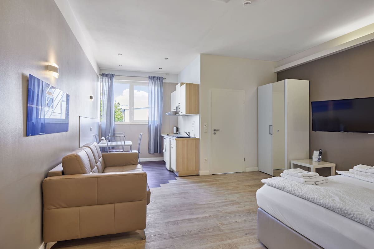 Habitación de hotel moderna con sofá, cama, televisor, zona de cocina y balcón con cortinas.