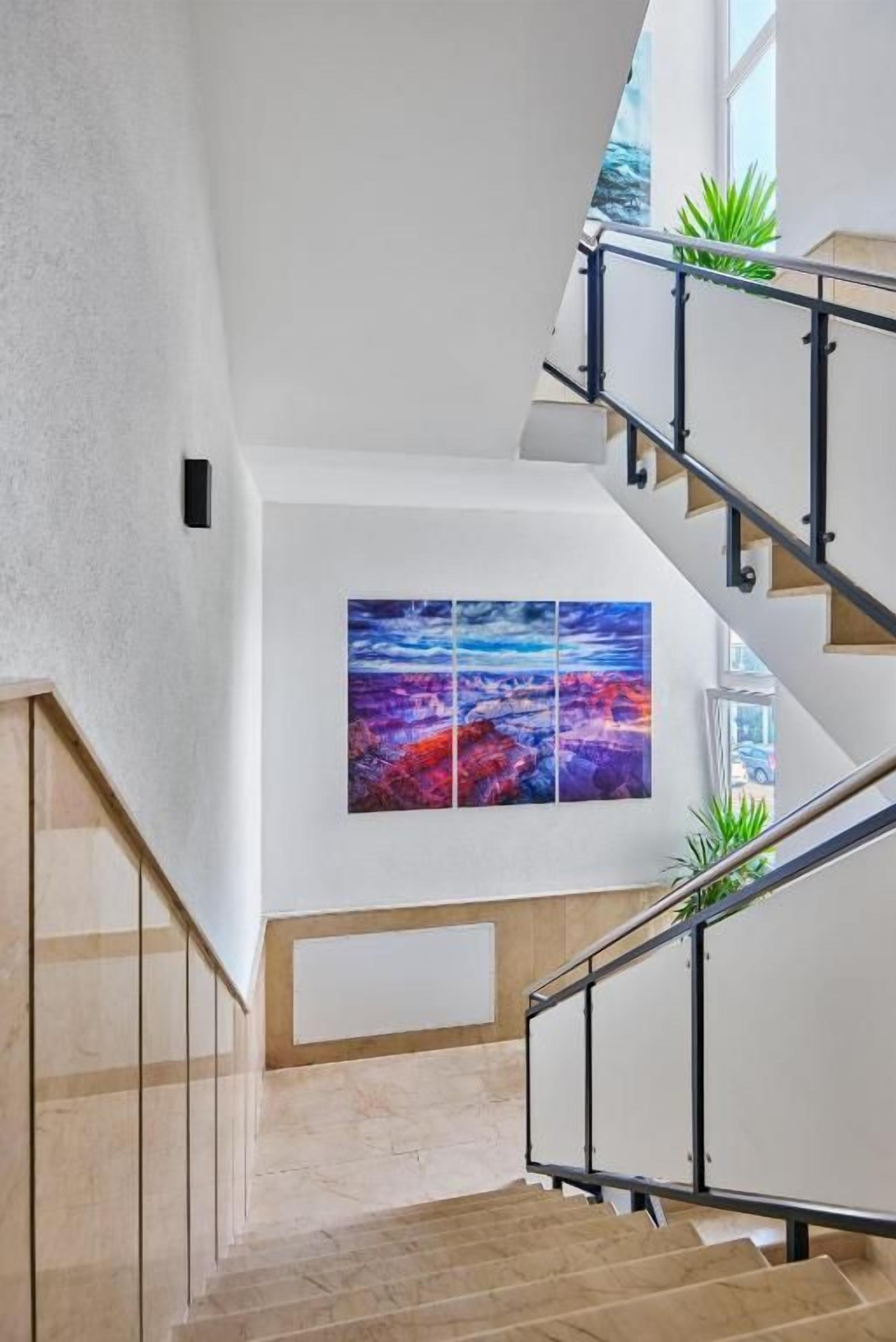 Uma escadaria moderna com corrimão de vidro, uma grande pintura abstrata na parede e um vaso de planta no patamar.