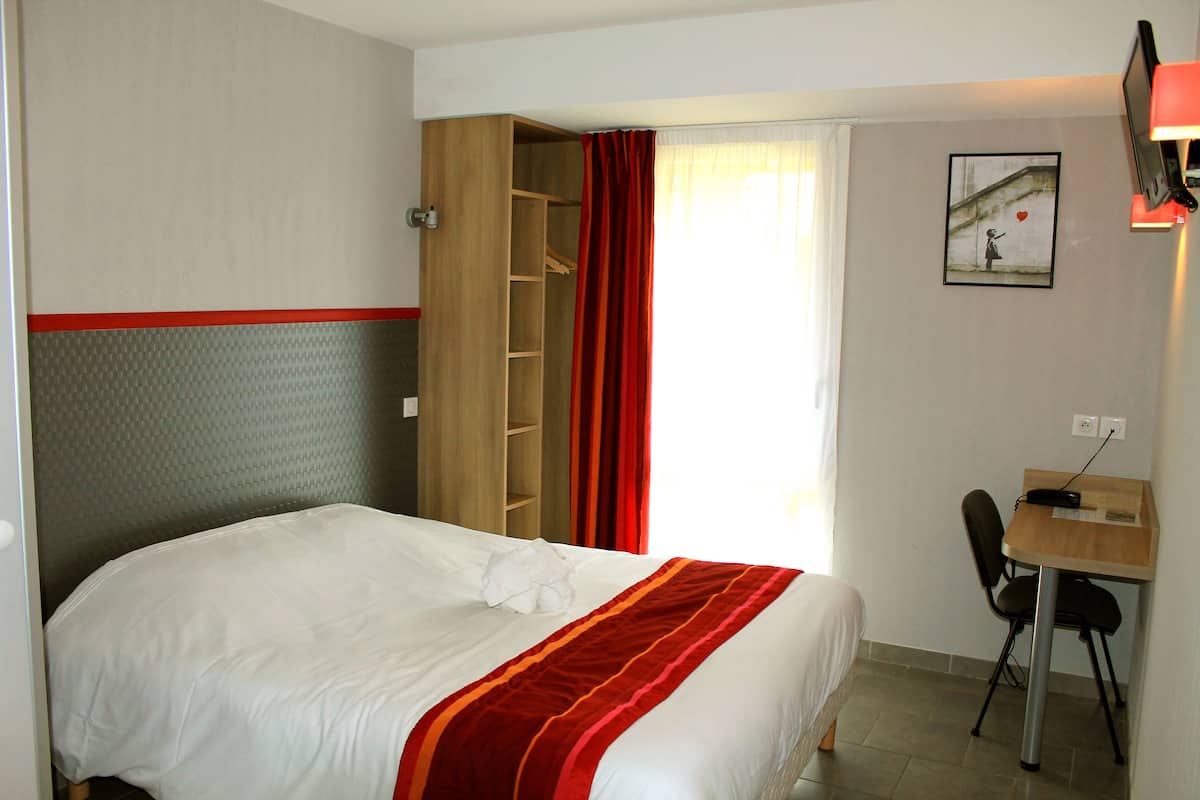 Una habitación de hotel con una cama, un escritorio y una silla. Se observa un armario con espejo y una cortina roja. Un cuadro adorna la pared.