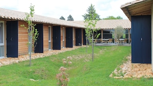 Een rij houten vakantiehuisjes met donkere luiken, een gazon en een terras met buitenmeubels.