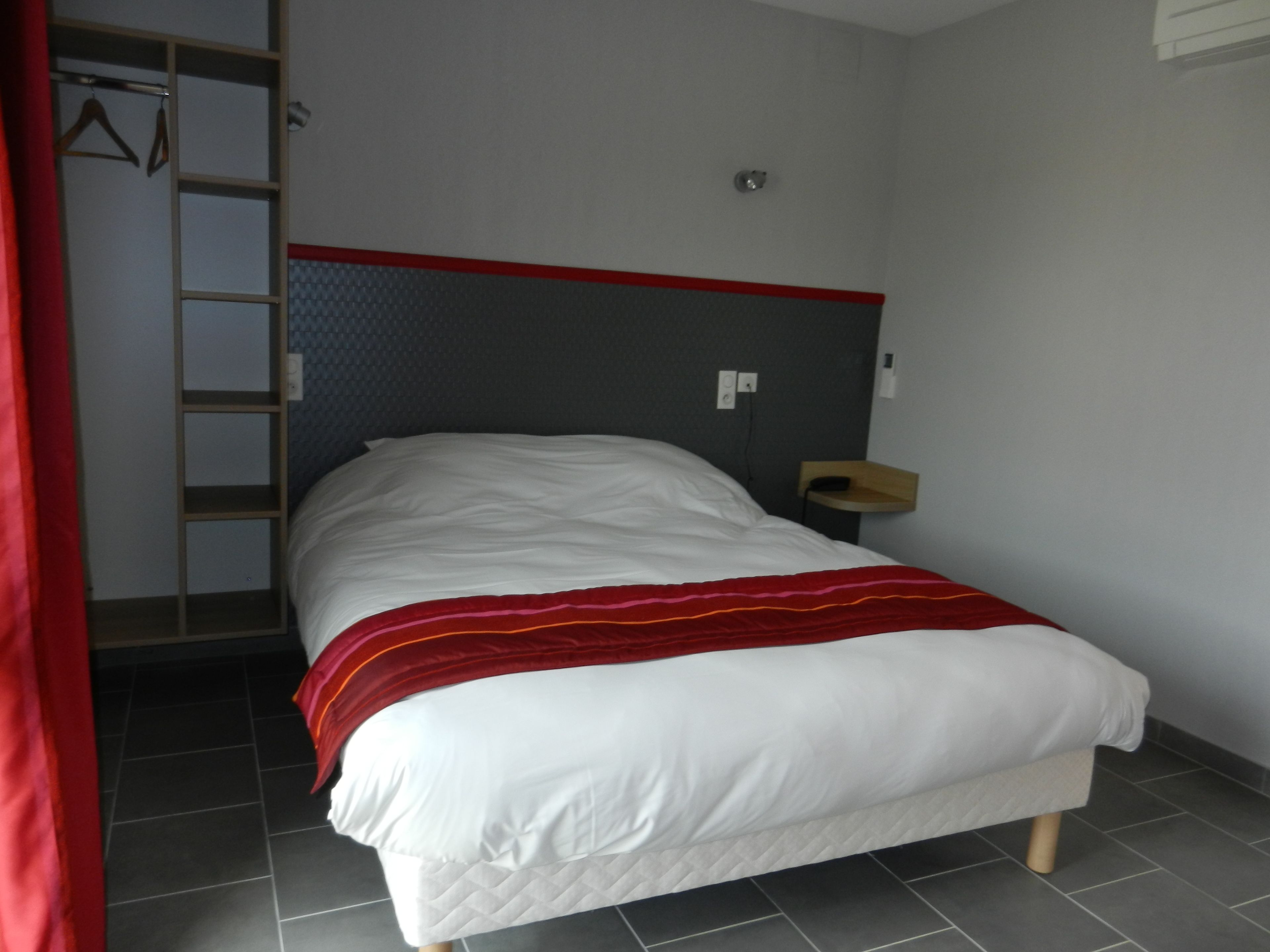 A neatly made bed with a red blanket, a wooden headboard, a bedside table, and a wall-mounted light.