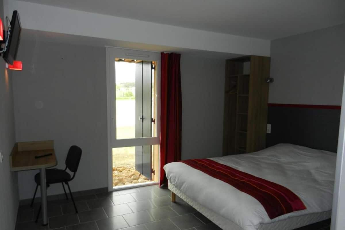 Un dormitorio con cama, escritorio y silla. Hay una puerta que da a un espacio exterior.