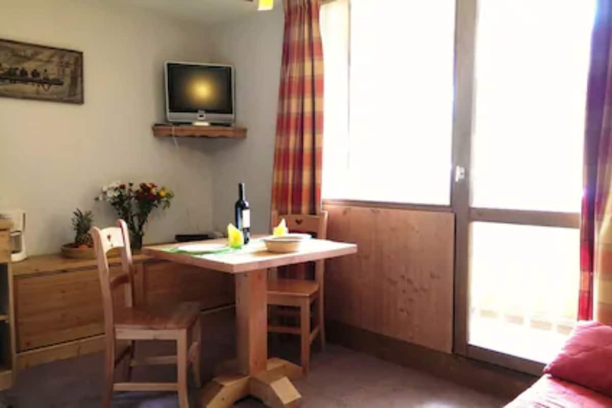 A room with a wooden desk, a chair, a television on a shelf, a window with curtains, and a bed with a red blanket.