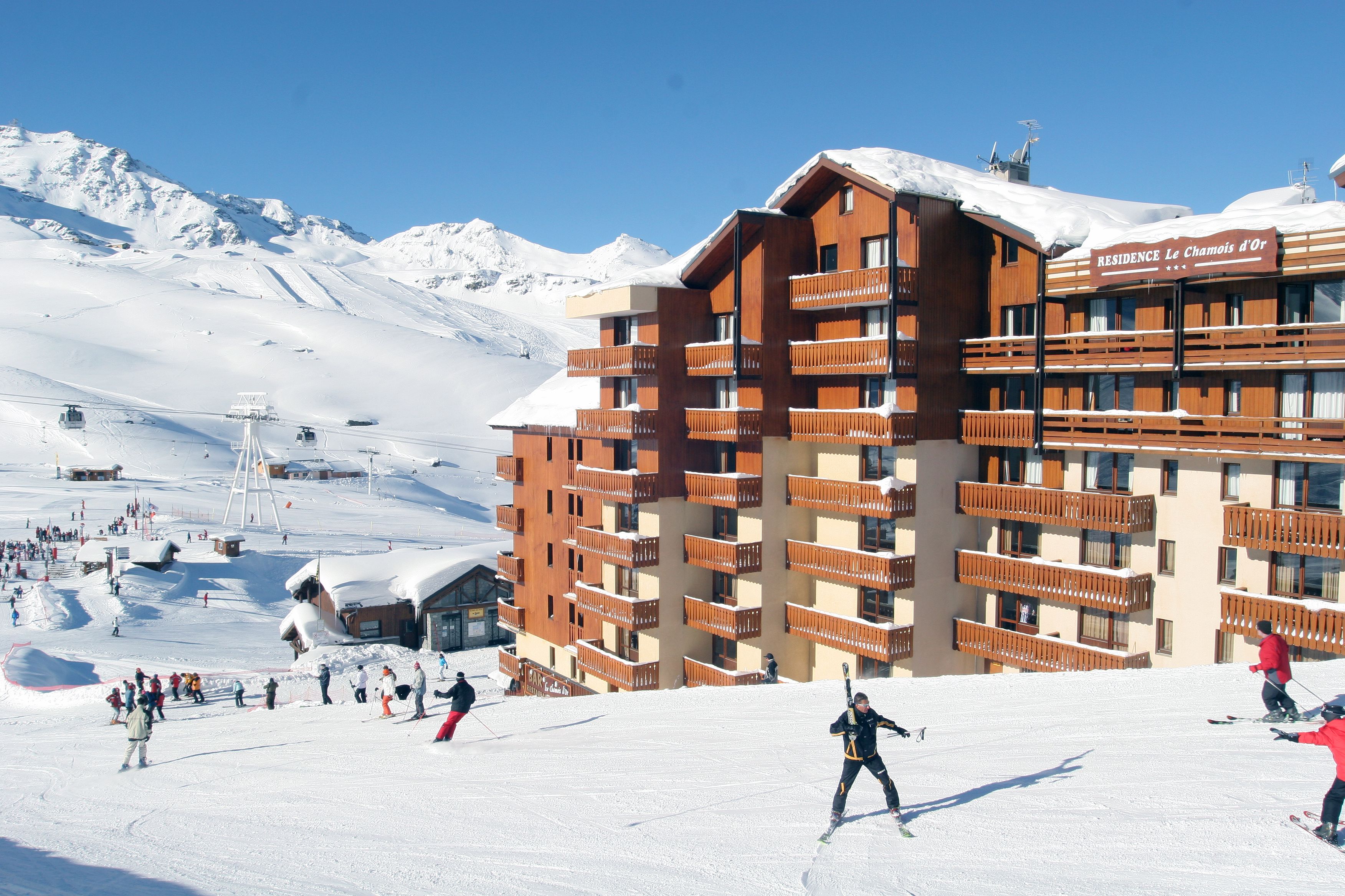 Un hotel de varios pisos con balcones, pistas de esquí al fondo y un cielo despejado.