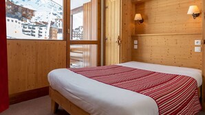 Una habitación de madera con una cama, dos lámparas fijadas a la pared y una ventana con vista a edificios.
