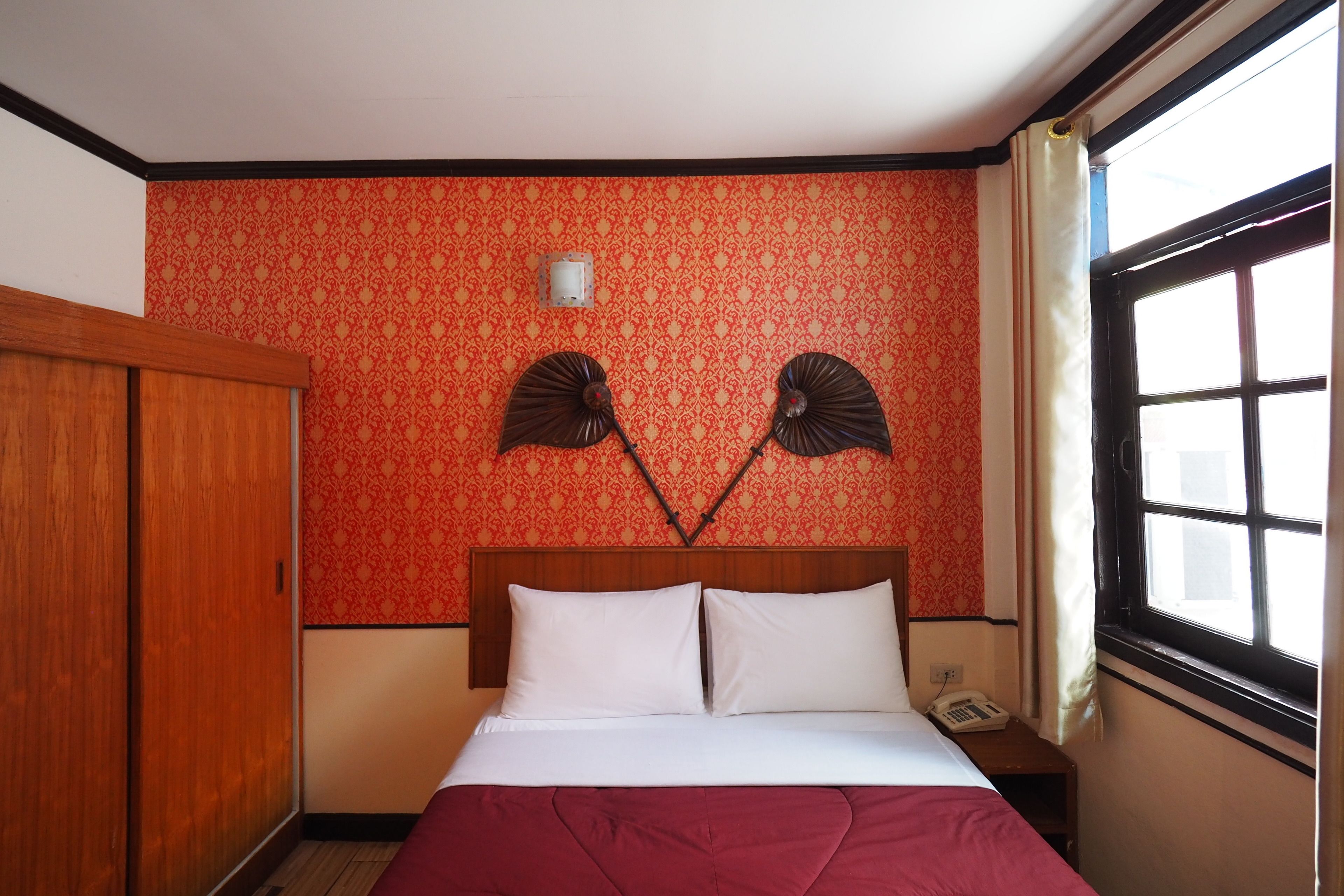 A hotel room with a bed, a wooden wardrobe, a wall-mounted phone, and a window with curtains.