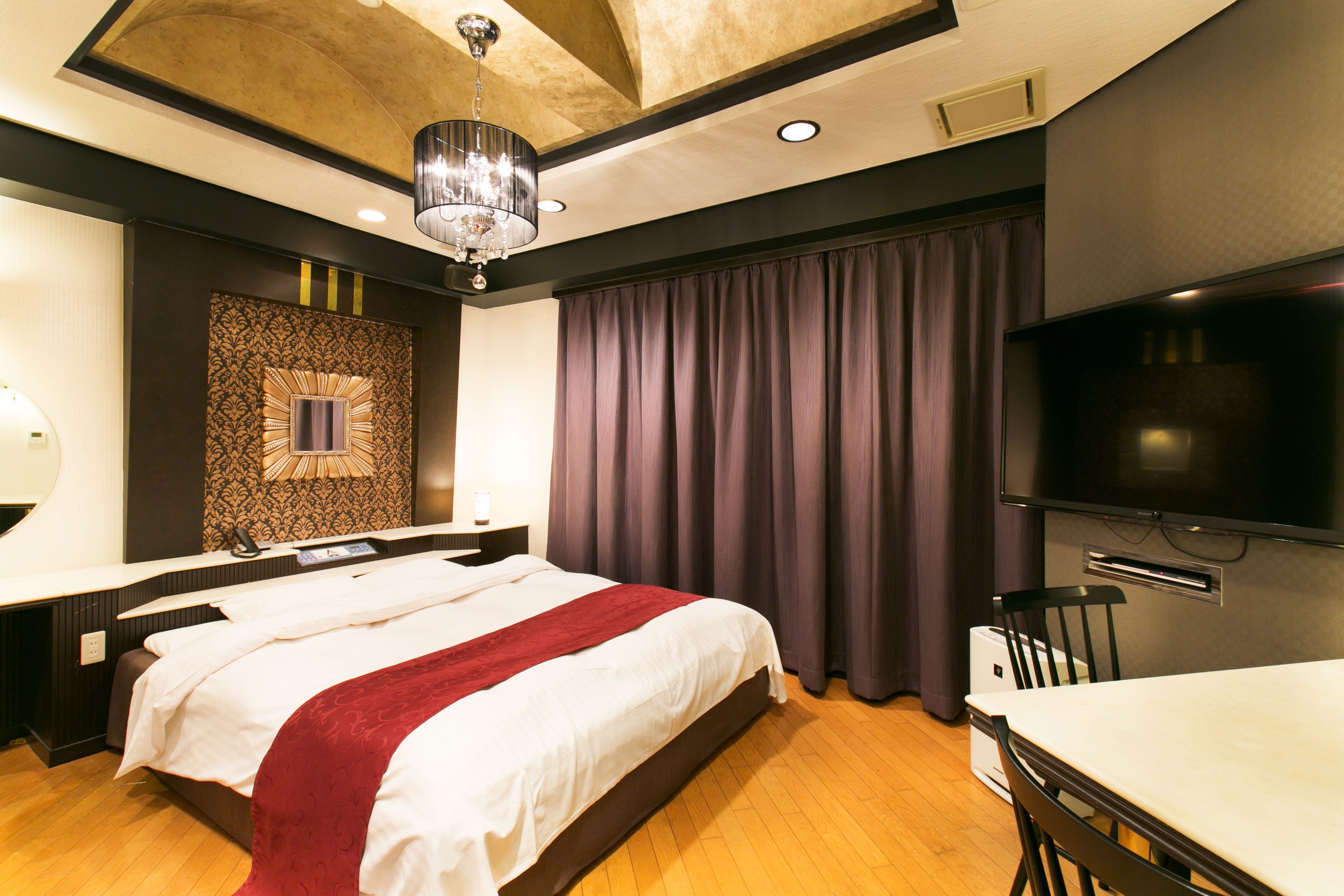 A modern hotel room with a large bed, a flat-screen TV, a desk with a chair, and a chandelier.