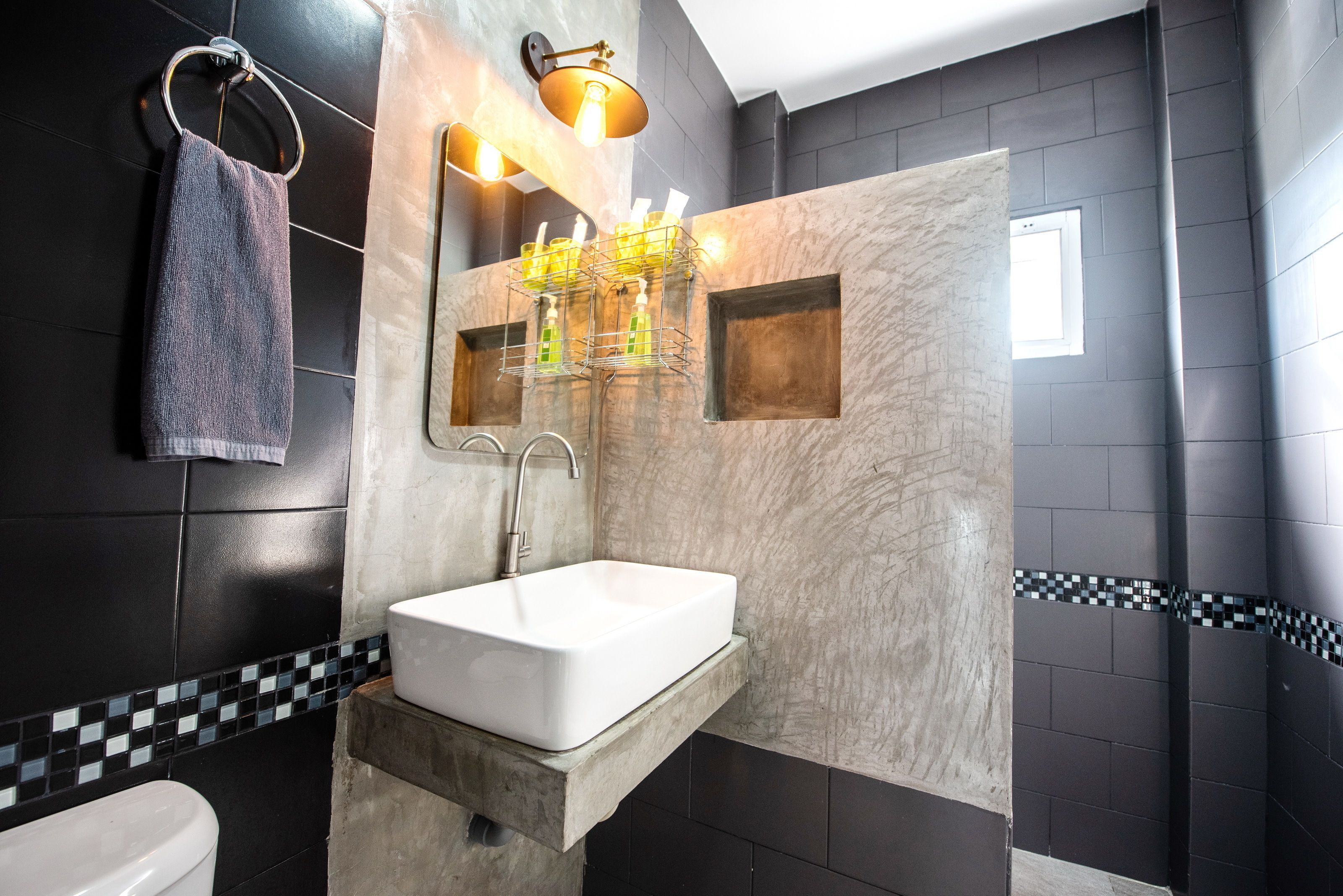 A modern bathroom with a white sink, dark tiled walls, a towel rack, and a mirror with lights.
