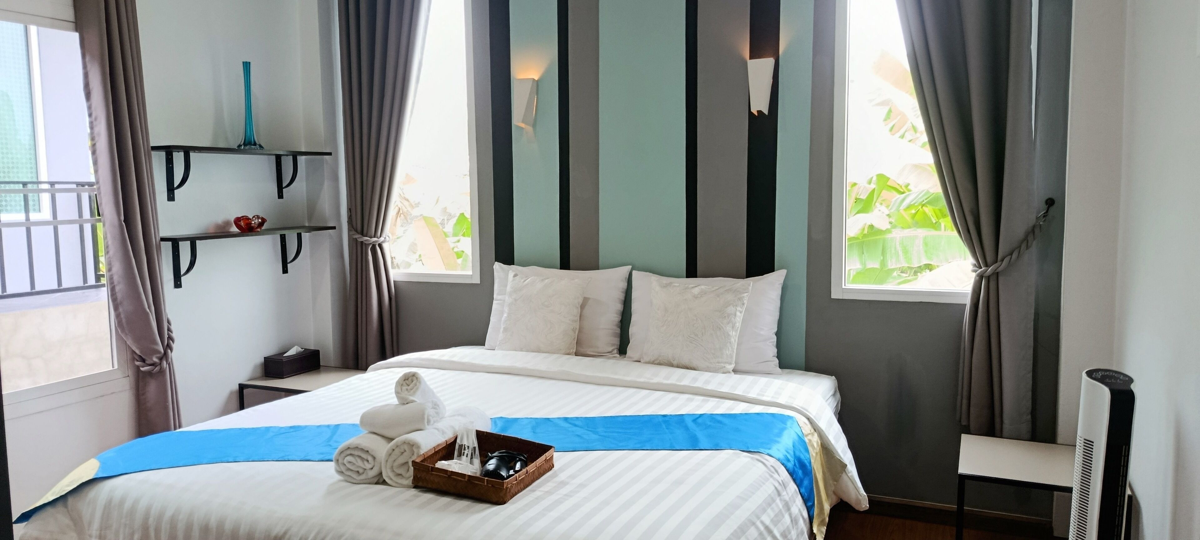 A neatly made bed with white linens and a blue accent, a bedside table with a lamp, and a window with curtains.