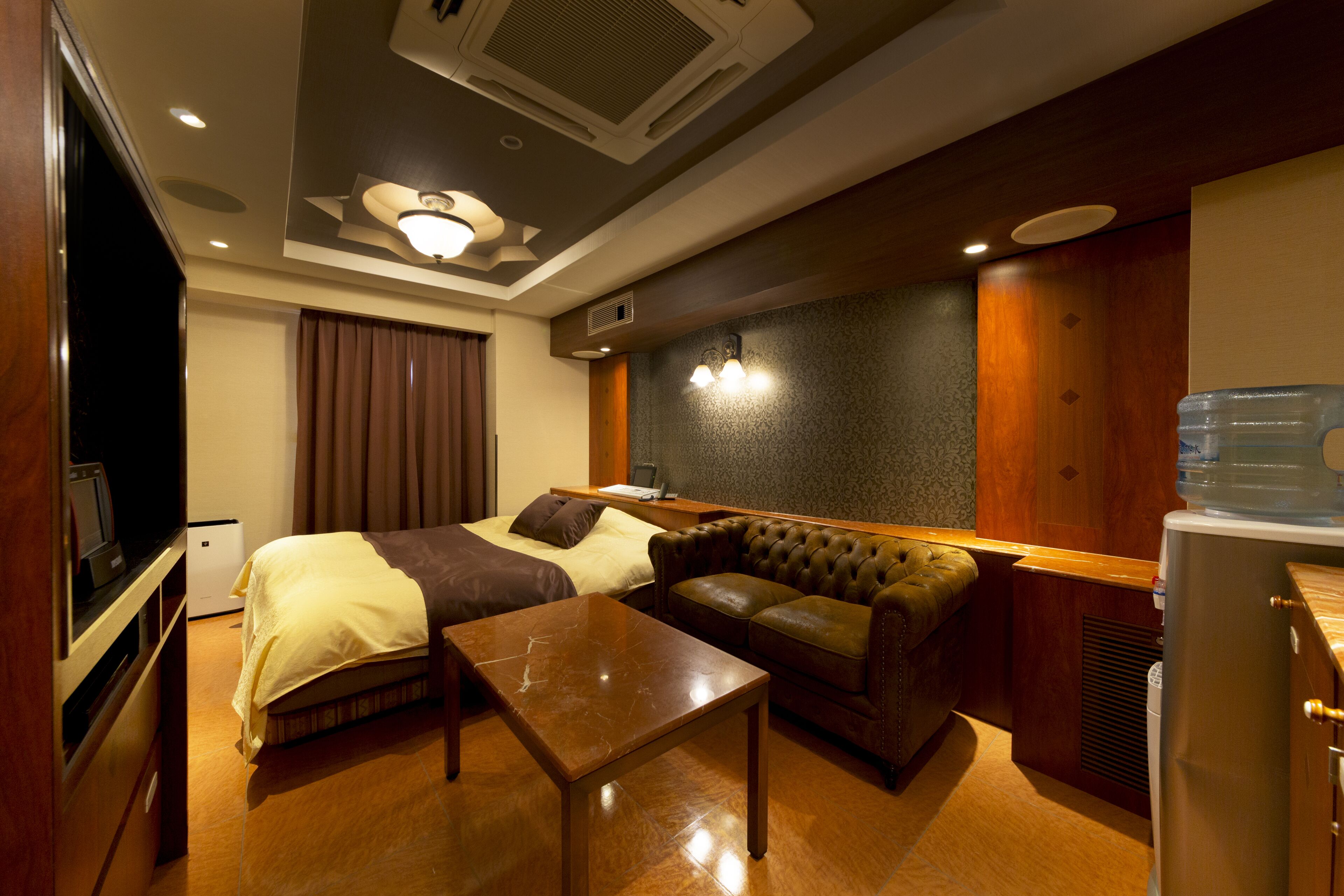 A hotel room with a bed, a leather sofa, a wooden coffee table, a flat-screen TV, a water dispenser, and a ceiling fan.