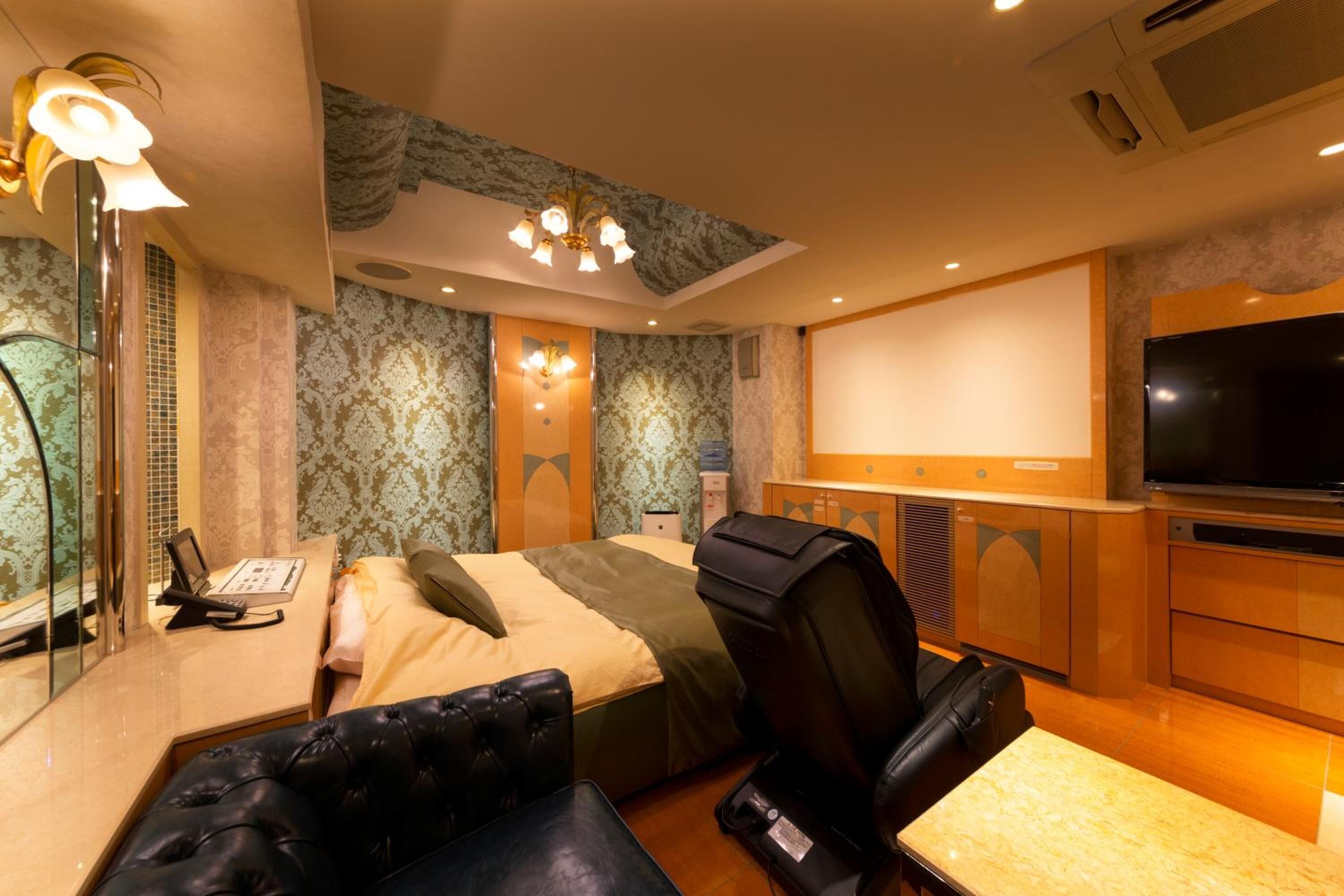 A hotel room with a bed, a massage chair, a desk with a phone, a TV, and a minibar.