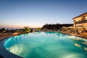 Ein Swimmingpool mit klarem, blauem Wasser, umgeben von Liegestühlen und Sonnenschirmen, mit einem Gebäude mit Balkon im Hintergrund.