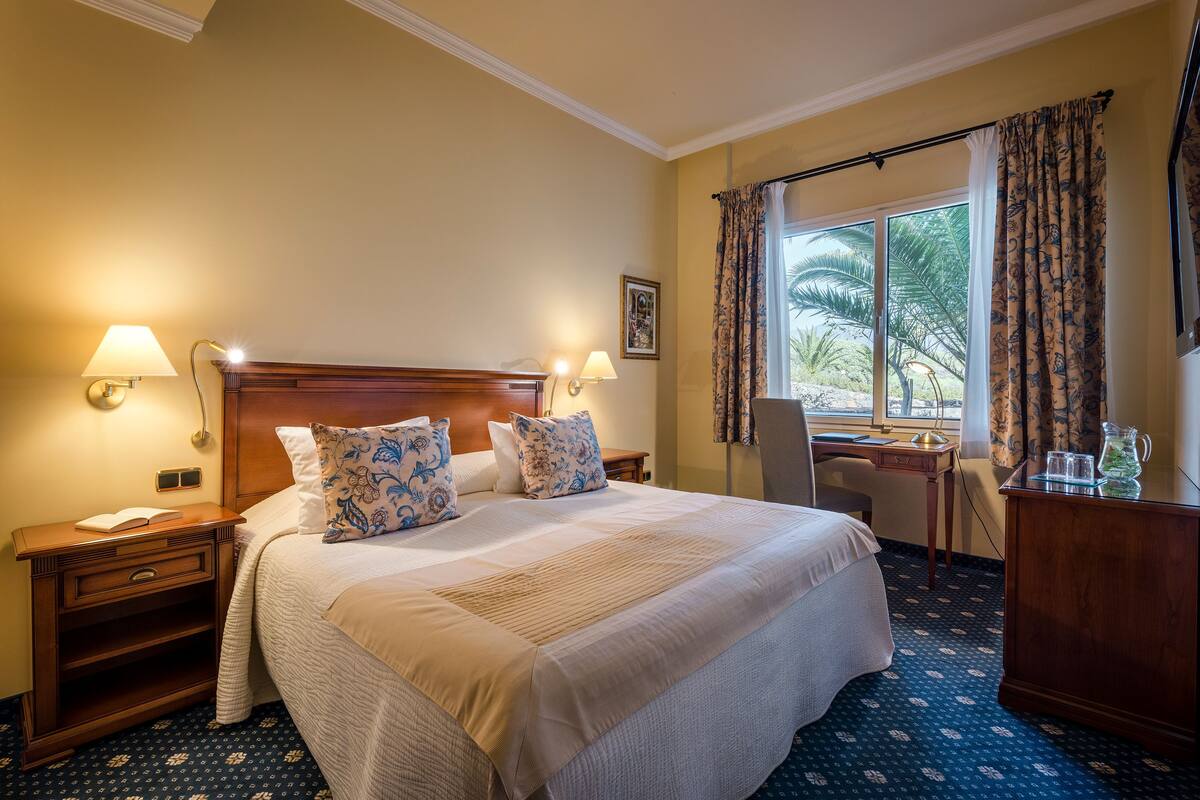 Una habitación de hotel con una cama grande, dos lámparas de noche, un escritorio y una ventana con cortinas.