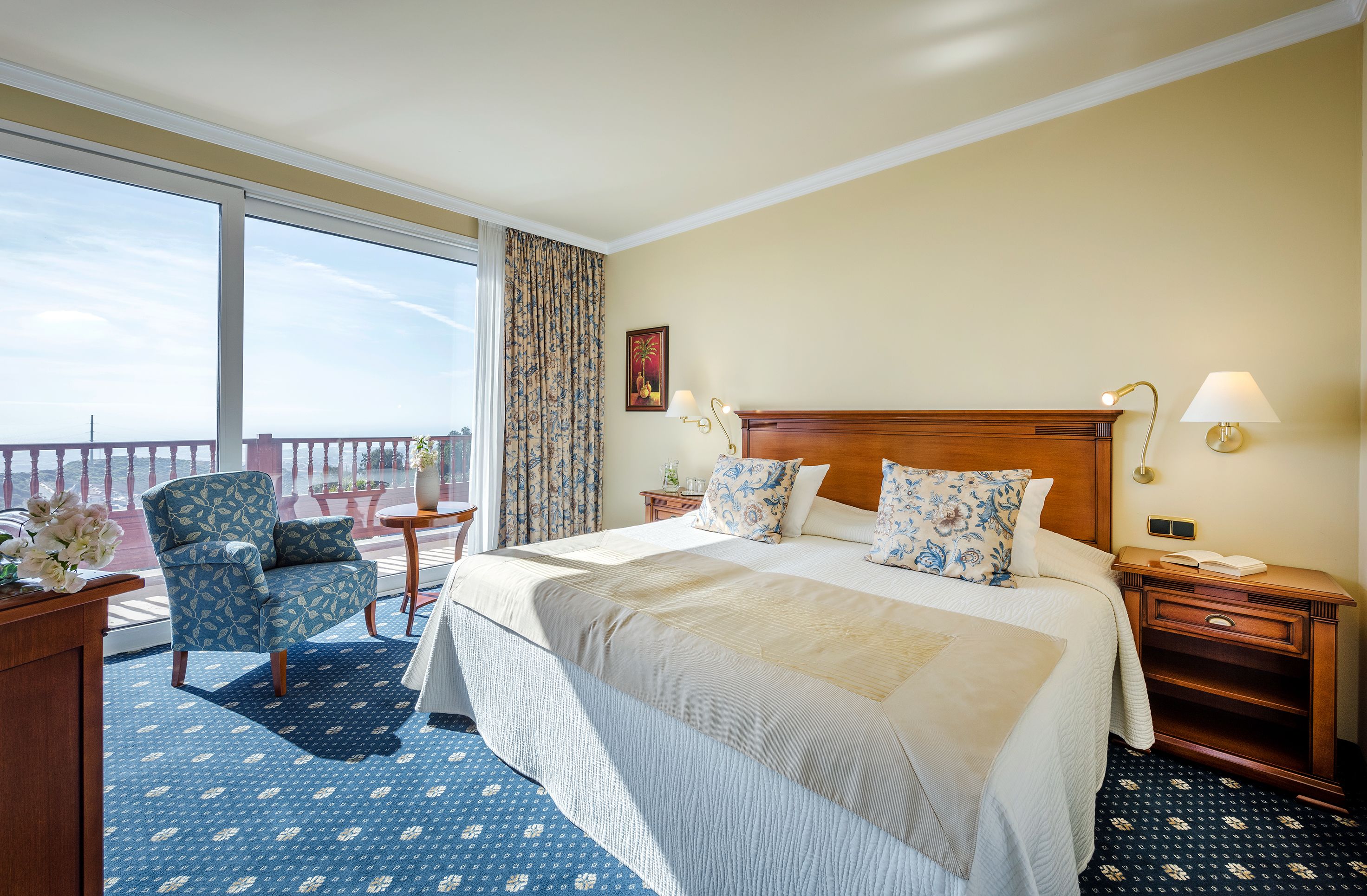 Habitación de hotel con una cama grande, una silla, una mesita de noche, una lámpara y vistas al exterior.