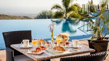 Desayuno con croissants, huevos y jugo en una mesa, cerca de una piscina con palmeras al fondo.