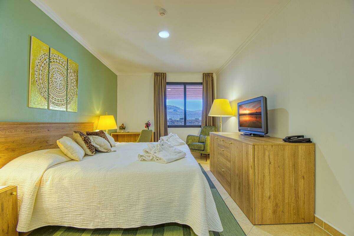Habitación de hotel con una cama grande, un aparador de madera, un televisor y vistas a la montaña desde la ventana.