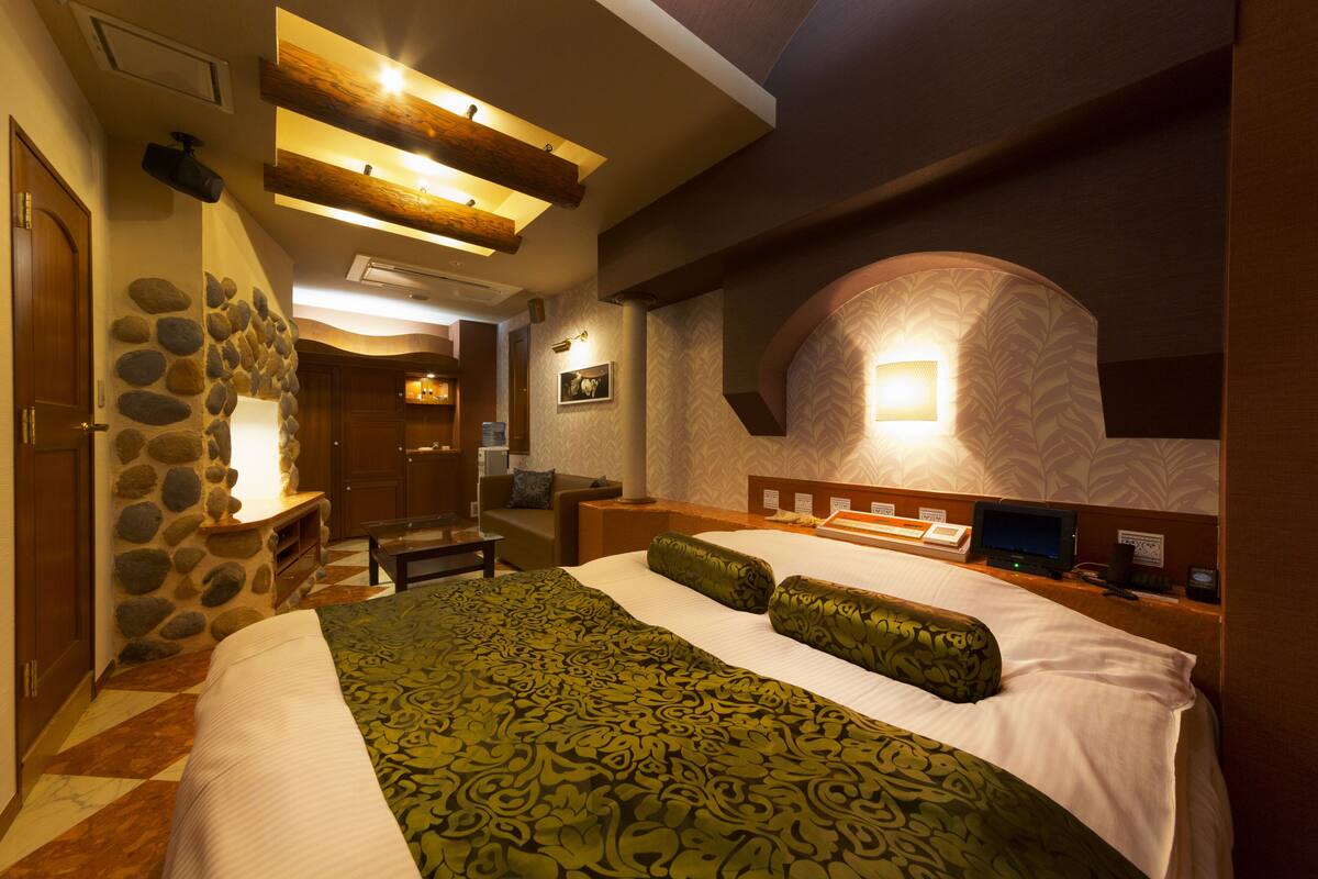 A hotel room with a large bed, a stone wall, a wooden ceiling, and a built-in desk with a computer.