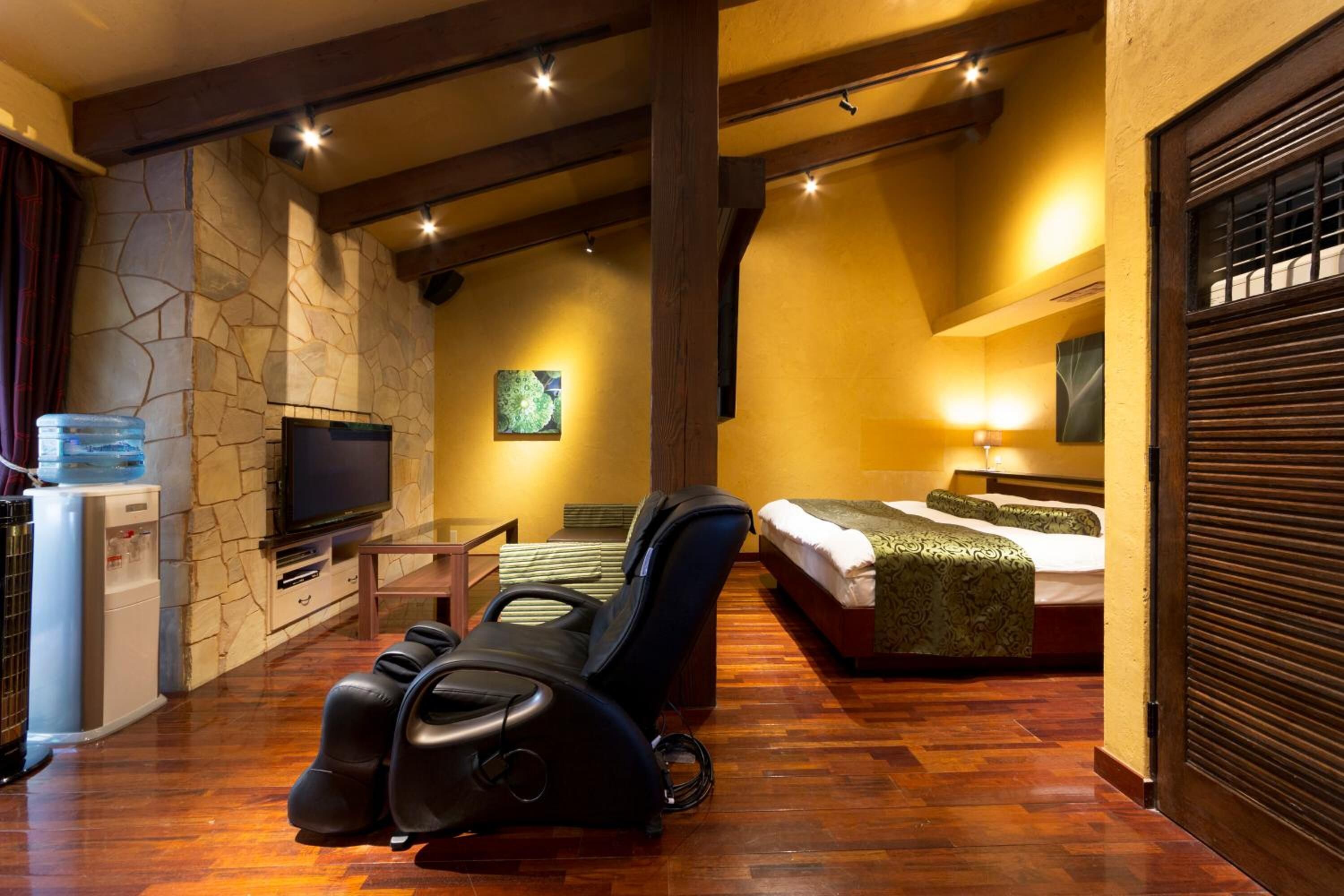 A room with a stone wall, a television, a massage chair, a bed with a green blanket, and a water dispenser.