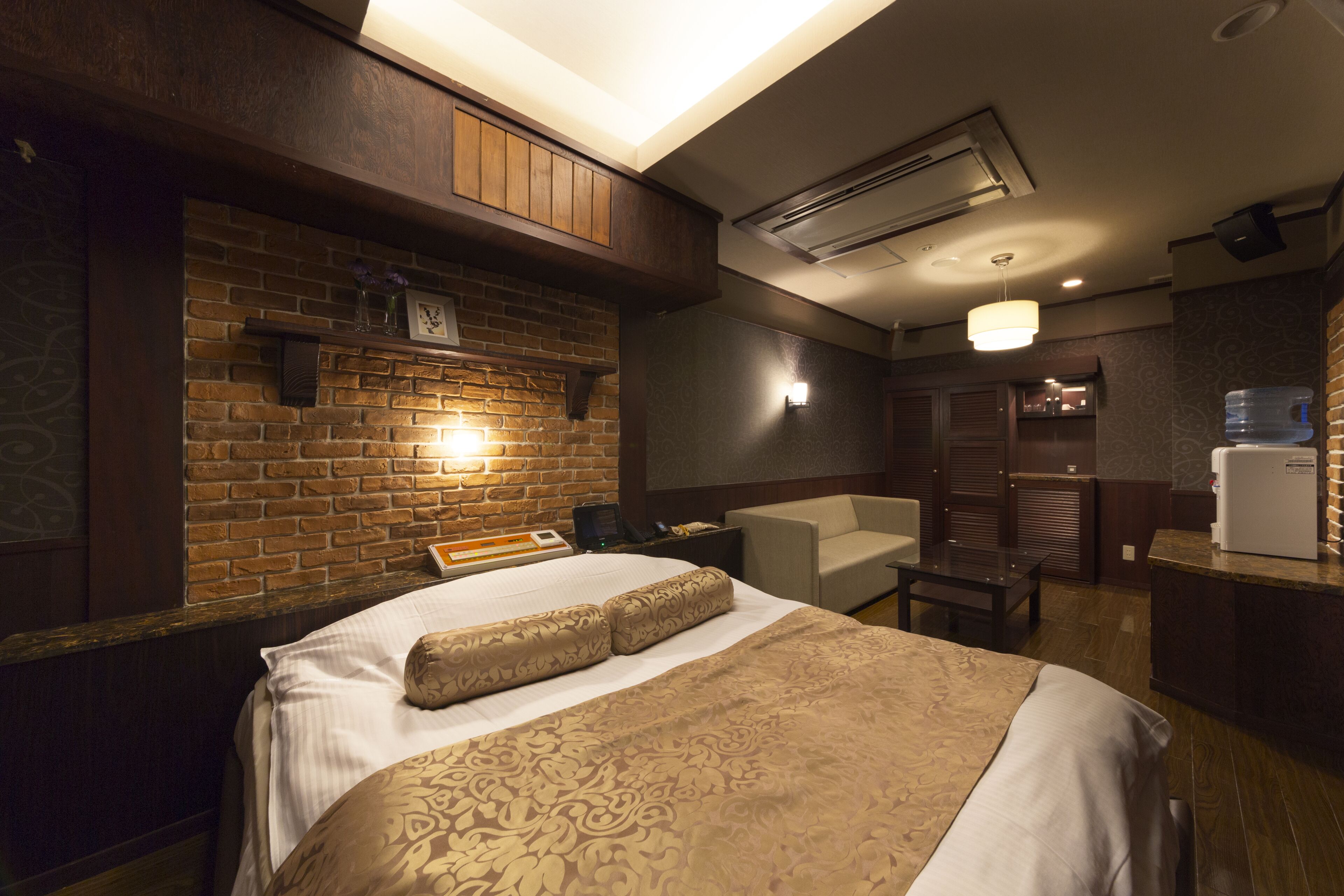 A hotel room with a brick wall, a bed with patterned bedding, a small table, a chair, and a water dispenser.
